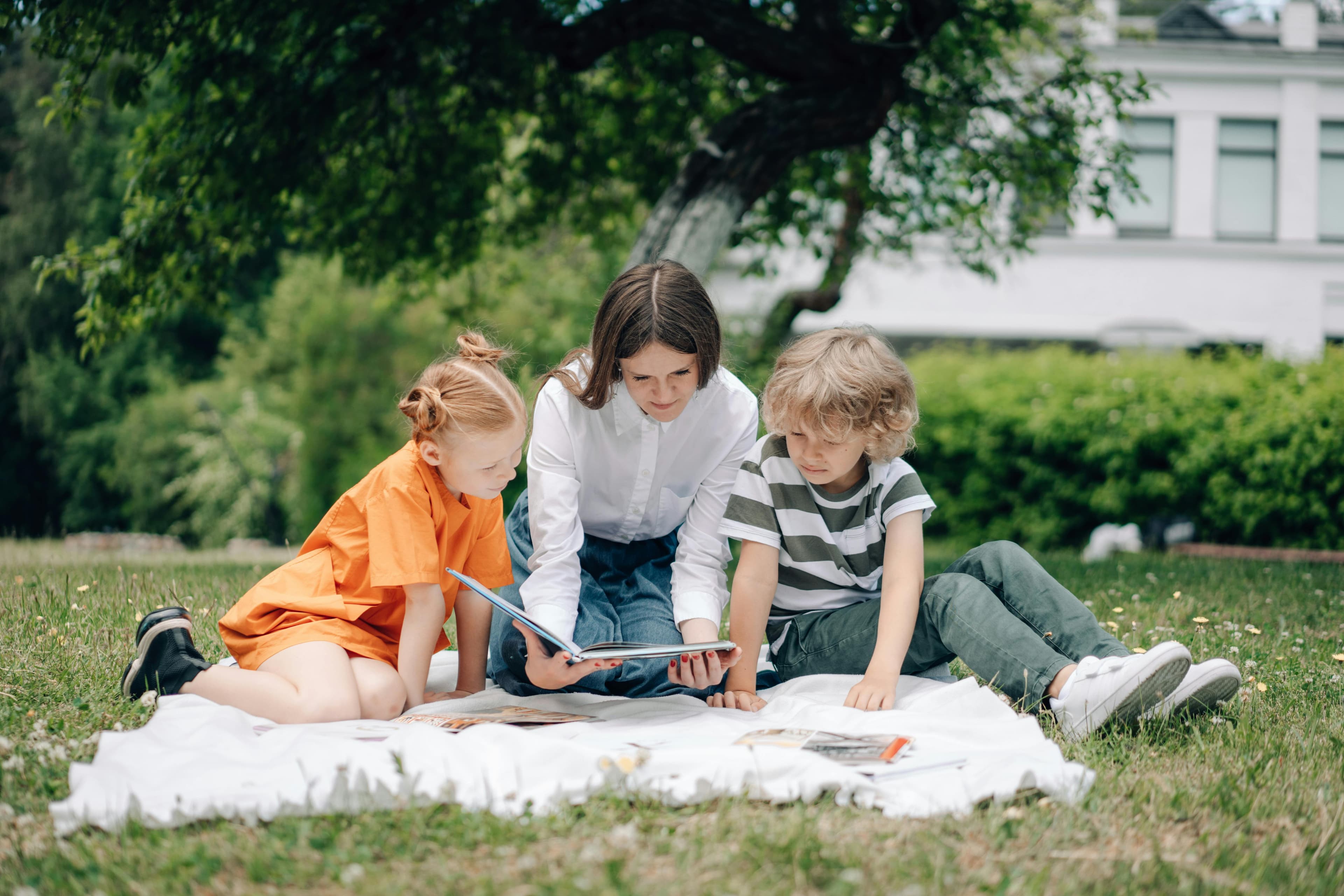 Erwachsene Person liest mit zwei Kindern gemeinsam ein Buch auf einer Decke im Park