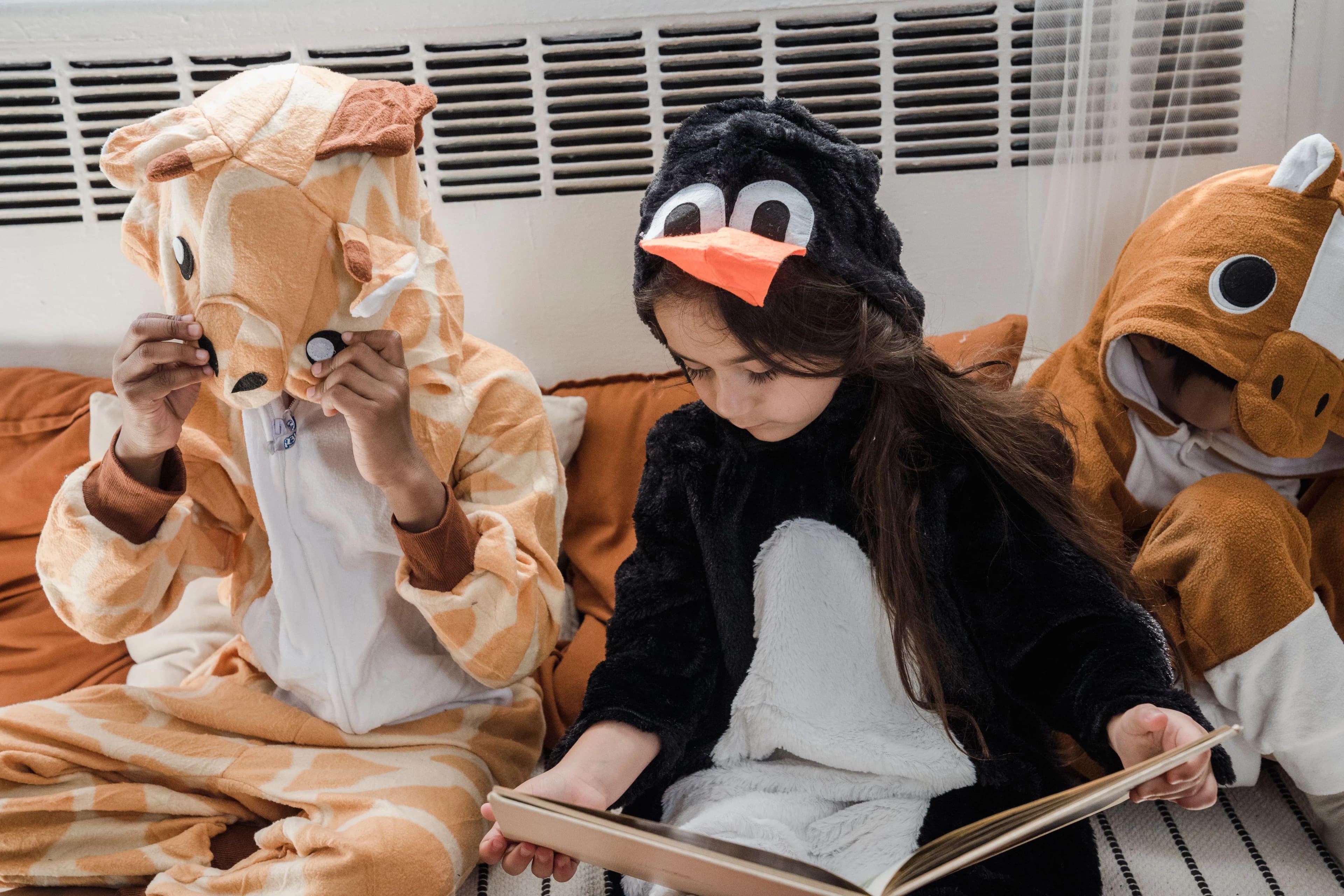 Kinder in Giraffen-, Pinguin- und Nashornkostümen sitzen auf einem Sofa und lesen gemeinsam ein Buch.